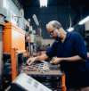 Factory worker with safety glasses and a beard operating a machine press, focused on precision work